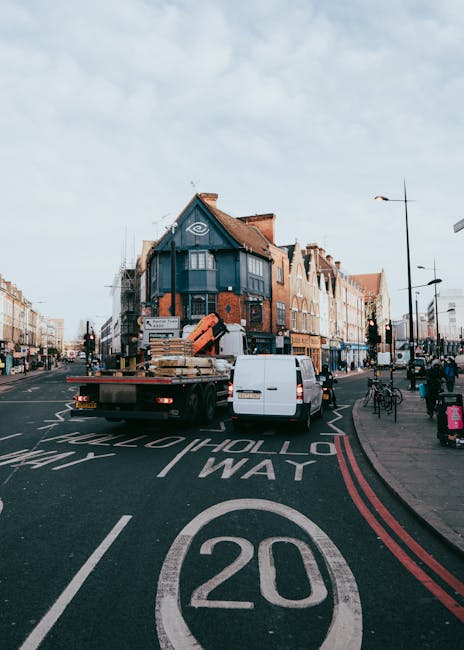A flatbed removal truck from Man with Van Muswell Hill is parked on Muswell Hill Broadway in London, close to a row of mixed-use buildings. The truck's flatbed is loaded with wooden pallets, cardboard boxes, and wrapped furniture, indicating a home relocation process. The vehicle is positioned partially inside a designated loading area marked by white lines, with the rear of the truck facing the street. A white van is parked beside the truck, assisting the loading or unloading activity. The pavement on the right side features bicycles and pedestrians, along with street lamps and signage. The buildings in the background are a mix of brick and painted facades, with windows and awnings, typical of urban residential and commercial properties. The road surface shows clear markings including a 20mph speed limit and directional arrows, contributing to the organized traffic flow for moving services conducted by [COMPANY_NAME]. The scene captures a typical moving or furniture transport scene, emphasizing the logistics involved in residential removals within the area.
