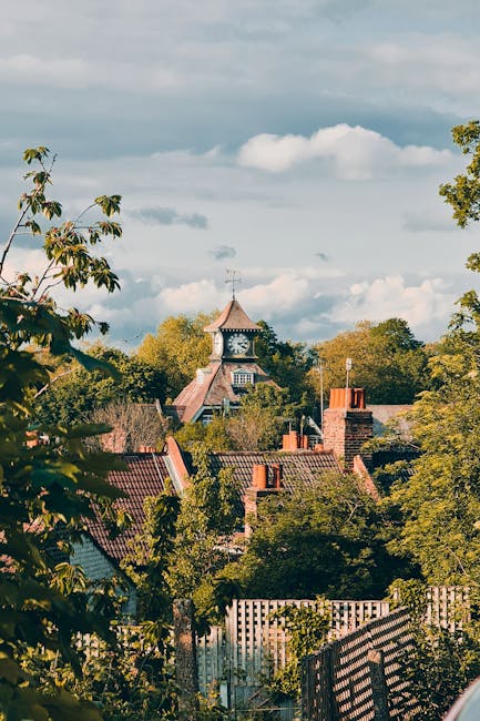 A view of a residential area in Muswell Hill with rooftops and chimneys visible among lush green trees under a partly cloudy sky. In the background, a historic clock tower with a pointed roof and weather vane stands prominently above the surrounding buildings. The scene captures the peaceful atmosphere typical of a home relocation zone, with no visible furniture or moving equipment. This image reflects the scenic environment where [COMPANY_NAME], such as Man with Van Muswell Hill, may carry out home and furniture transport. The natural daylight highlights the textures of the rooftops, chimney stacks, and the clock tower, emphasizing the peaceful yet busy context of moving and packing services within a picturesque London suburb.