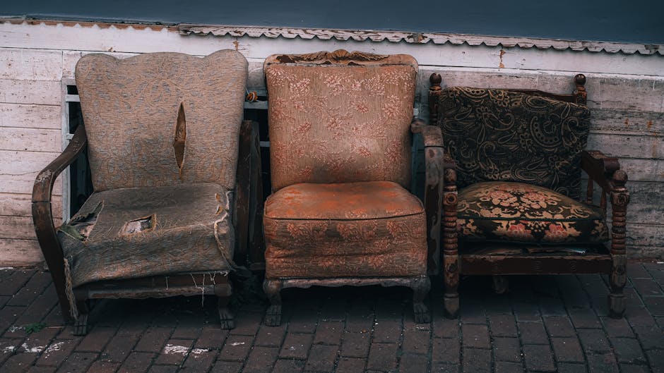 A row of three antique upholstered armchairs with wooden frames and decorative fabric, positioned outside a weathered white wooden building wall on a brick pavement. The left armchair shows significant wear with torn upholstery and exposed padding, the middle armchair has faded, patterned fabric with some visible stains, and the right armchair features dark, patterned fabric with intact cushioning. The setting appears to be part of a house removal process, with these chairs likely prepared for disposal or transport, as part of furniture removals carried out by Man with Van Muswell Hill. The scene reflects an outdoor area where furniture is being loaded or unloaded, with attention to the condition and placement of each piece in a professional move involving packing, lifting, or disposal of bulky furniture during the home relocation steps.
