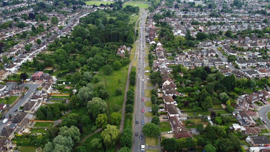 An aerial view of a residential street in Muswell Hill showing a narrow street lined with parked cars and front gardens, adjacent to a green wooded area with tall trees and open grassy patches. The street is flanked by houses with pitched roofs, some with visible driveways and gardens, and the scene appears to be taken during daylight hours with clear visibility. This snapshot of a typical neighborhood on Colney Hatch Lane highlights the residential environment and urban greenery, suitable for illustrating home relocation or furniture transport services, with the image capturing the calm, tree-lined street ideal for moving logistics by a professional removals company like Man with Van Muswell Hill.