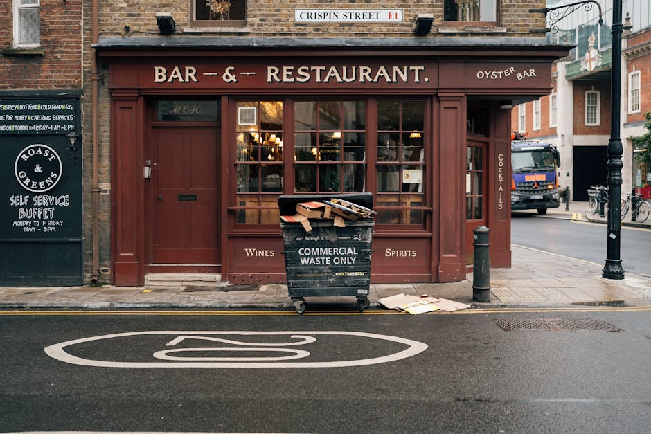 The image shows the front exterior of a small restaurant or bar located on a city street, with large windows revealing a warmly lit interior. The establishment's sign above the windows displays 'BAR &