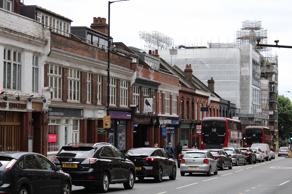 A street view of residential and commercial buildings with brick facades and large sash windows in Muswell Hill, Haringey. Several cars, including black, grey, and silver vehicles, are parked along the pavement, with some moving traffic visible. Two red double-decker buses, numbered 343 and 142, are traveling down the road amidst the parked cars. The scene includes construction scaffolding on a building to the right, covered with white sheeting, indicating ongoing refurbishment. The pavement is occupied by pedestrians, some walking and others possibly organizing belongings for home relocation. Visible elements such as cardboard boxes, packing materials, and furniture being loaded or unloaded are not present in this image; however, the environment illustrates typical urban moving logistics. Occasionally, Man with Van Muswell Hill offers professional removals and furniture transport services, often involved in busy residential or commercial moving activities within the area.