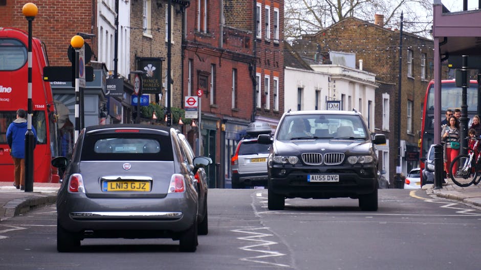 A street view of residential and commercial buildings with brick facades and large sash windows in Muswell Hill, Haringey. Several cars, including black, grey, and silver vehicles, are parked along the pavement, with some moving traffic visible. Two red double-decker buses, numbered 343 and 142, are traveling down the road amidst the parked cars. The scene includes construction scaffolding on a building to the right, covered with white sheeting, indicating ongoing refurbishment. The pavement is occupied by pedestrians, some walking and others possibly organizing belongings for home relocation. Visible elements such as cardboard boxes, packing materials, and furniture being loaded or unloaded are not present in this image; however, the environment illustrates typical urban moving logistics. Occasionally, Man with Van Muswell Hill offers professional removals and furniture transport services, often involved in busy residential or commercial moving activities within the area.