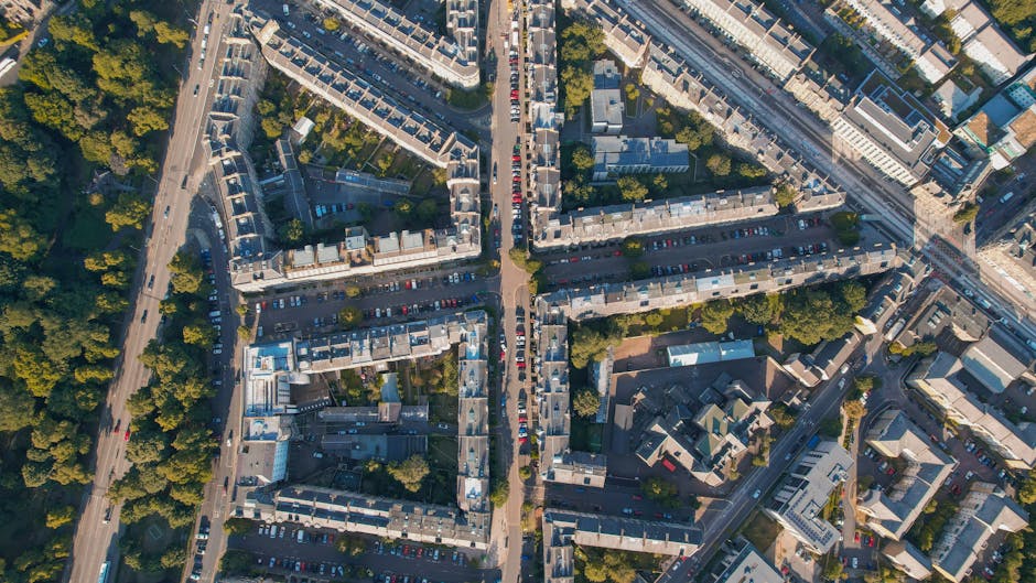 Aerial view of a densely built urban area showing multiple rows of terraced brick houses with grey rooftops, aligned along narrow streets. The streets are filled with parked cars, some walking paths, and occasional trees lining the pavements. To the left, a highway with multiple lanes is visible, bordered by a line of green trees. In the upper right corner, part of a modern multi-storey building is visible, with a large road passing nearby. The image captures the typical residential layout of a London neighbourhood, illustrating the setting where house removals or furniture transport may occur during local moves or packing and moving activities. The lighting indicates daytime with shadows cast by the buildings and trees, highlighting the urban environment. For house removals or home relocation services, Man with Van Muswell Hill operates in this area, providing efficient transport and logistical support around Alexandra Palace and Muswell Hill.