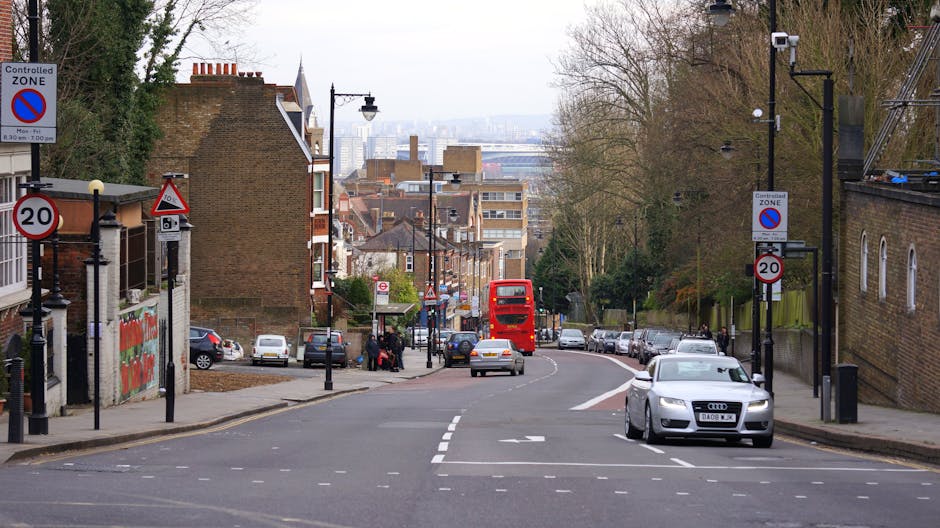 A street scene near Alexandra Palace in Muswell Hill shows a large, white, neoclassical-style building with columns and ornate detailing on the left side, partially obscured by leafless trees. In the background, the iconic dome of Alexandra Palace is visible behind the trees. The road curves gently to the right, with some pedestrians and cyclists moving along the pavement, which is protected by metal barriers. Several vehicles are parked along the street, and orange and blue traffic bollards mark the edge of the loading area. The scene is illuminated by natural daylight, with clear skies overhead. The image captures a quiet moment during housing or furniture transport activities, with an emphasis on urban surroundings suitable for house removals or moving services provided by Man with Van Muswell Hill, especially around restricted access areas like Alexandra Palace.