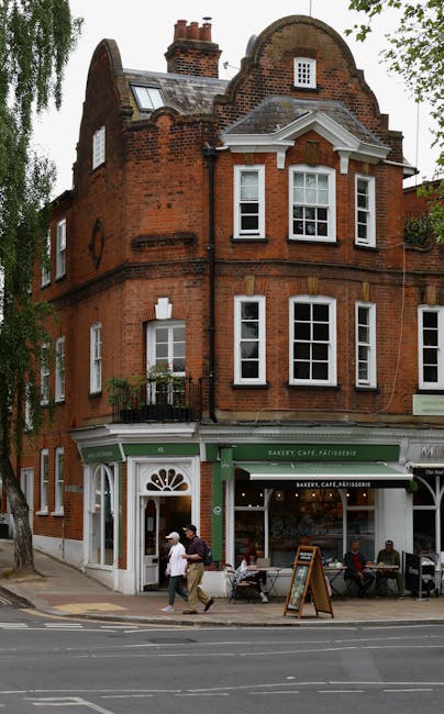 A three-storey red brick building situated on a busy street corner in Muswell Hill, with a bakery café on the ground floor featuring a green and white storefront. The café has a large window display and a white awning, with outdoor seating visible on the sidewalk, where several people are seated. Two pedestrians, one wearing a white shirt and the other in dark clothing, are walking past the entrance. The upper floors have multiple white-framed sash windows, some with small balcony railings. The building is surrounded by trees, and the street shows parked and moving vehicles, with a clear sky providing natural daylight for a daytime scene. This setting illustrates the typical architecture found in the area, relevant to house removals and moving services offered by Man with Van Muswell Hill as part of their home relocation and furniture transport operations.