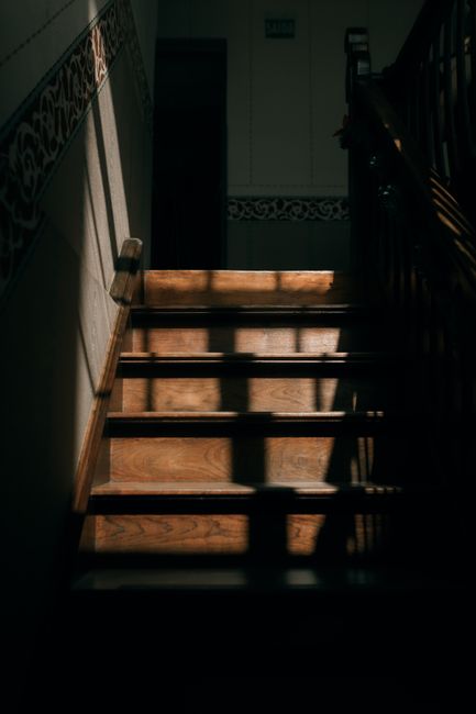 Inside a residential property showing a wooden staircase with dark handrails and decorative side panels, illuminated by natural light casting shadows on the steps. The staircase is part of a home undergoing a move, with no visible furniture or boxes on the stairs themselves. The surrounding walls are painted in a light colour, and the upward view highlights the elevation within the interior space. The scene suggests a focus on the staircase area during a home relocation process, with attention to the structural elements and lighting. Man with Van Muswell Hill’s removal services may involve managing furniture transport through this staircase, ensuring safe and efficient movement within the property.