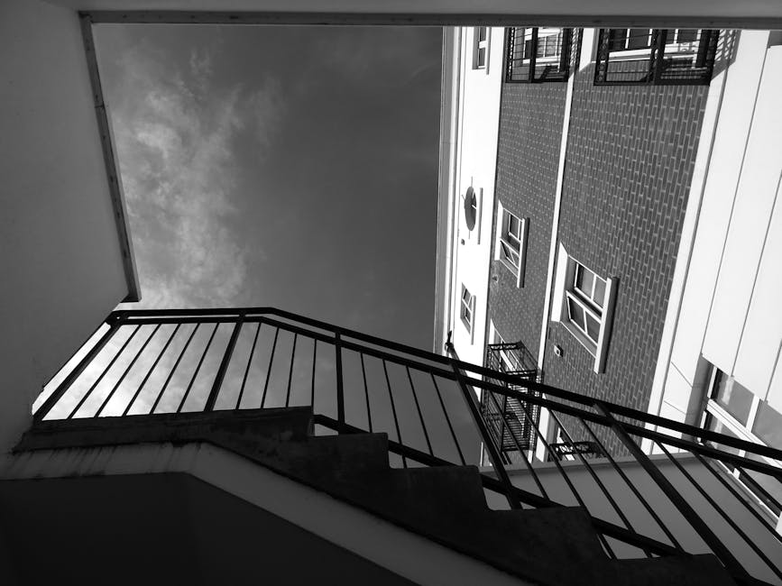 Black-and-white photograph capturing the exterior staircase of a staircase-only flat in Muswell Hill, showing metal railings and concrete steps leading to an upper level. The image is taken from below, looking upward toward the sky, and includes views of neighboring facades of residential buildings with brickwork and windows. The surroundings suggest an urban residential environment, with the staircase positioned inside an outdoor balcony or fire escape structure. The scene depicts a typical setup where furniture and boxes may be carried for home relocation or moving services, as part of house removals provided by Man with Van Muswell Hill. The natural lighting highlights the contrast between the dark metal railing and lighter building surfaces, emphasizing the practical environment involved in furniture transport or packing during the loading process for a safe and organized move.