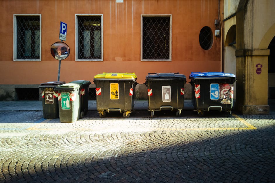A small commercial waste collection truck is parked on a street in Muswell Hill during daylight, with the rear hopper open and filled with various recycled materials such as cardboard boxes, plastic b